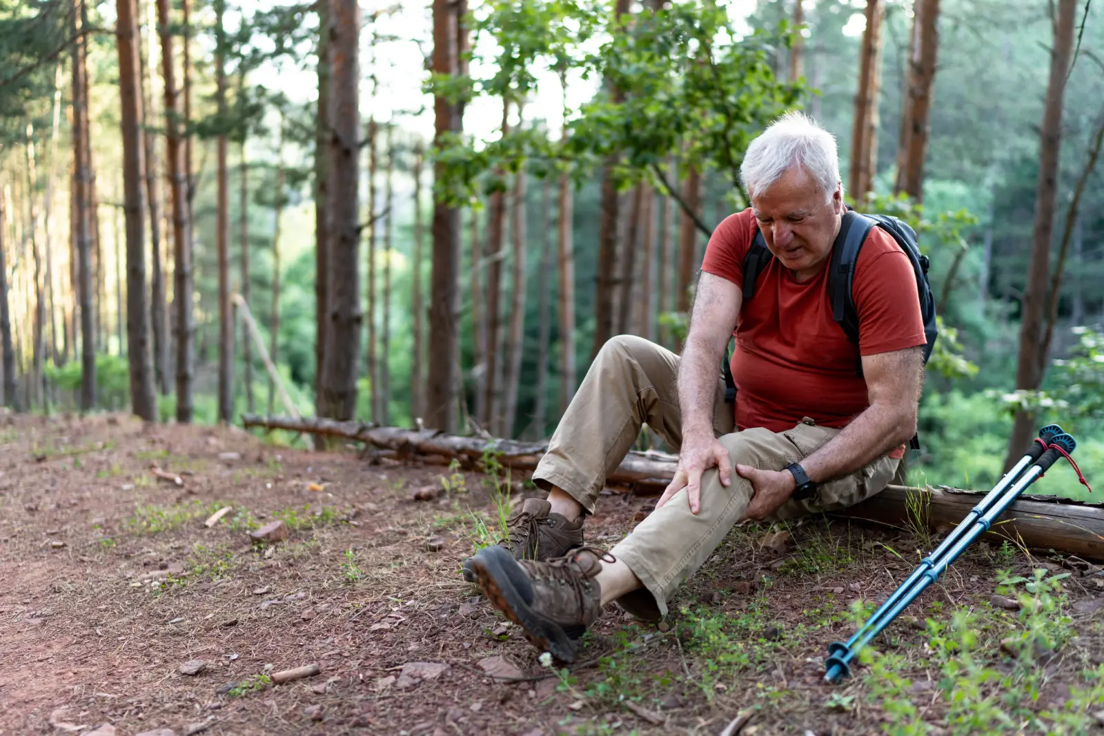 An older man in hiking gear sits beside a forest trail holding his leg, trekking poles resting beside him.